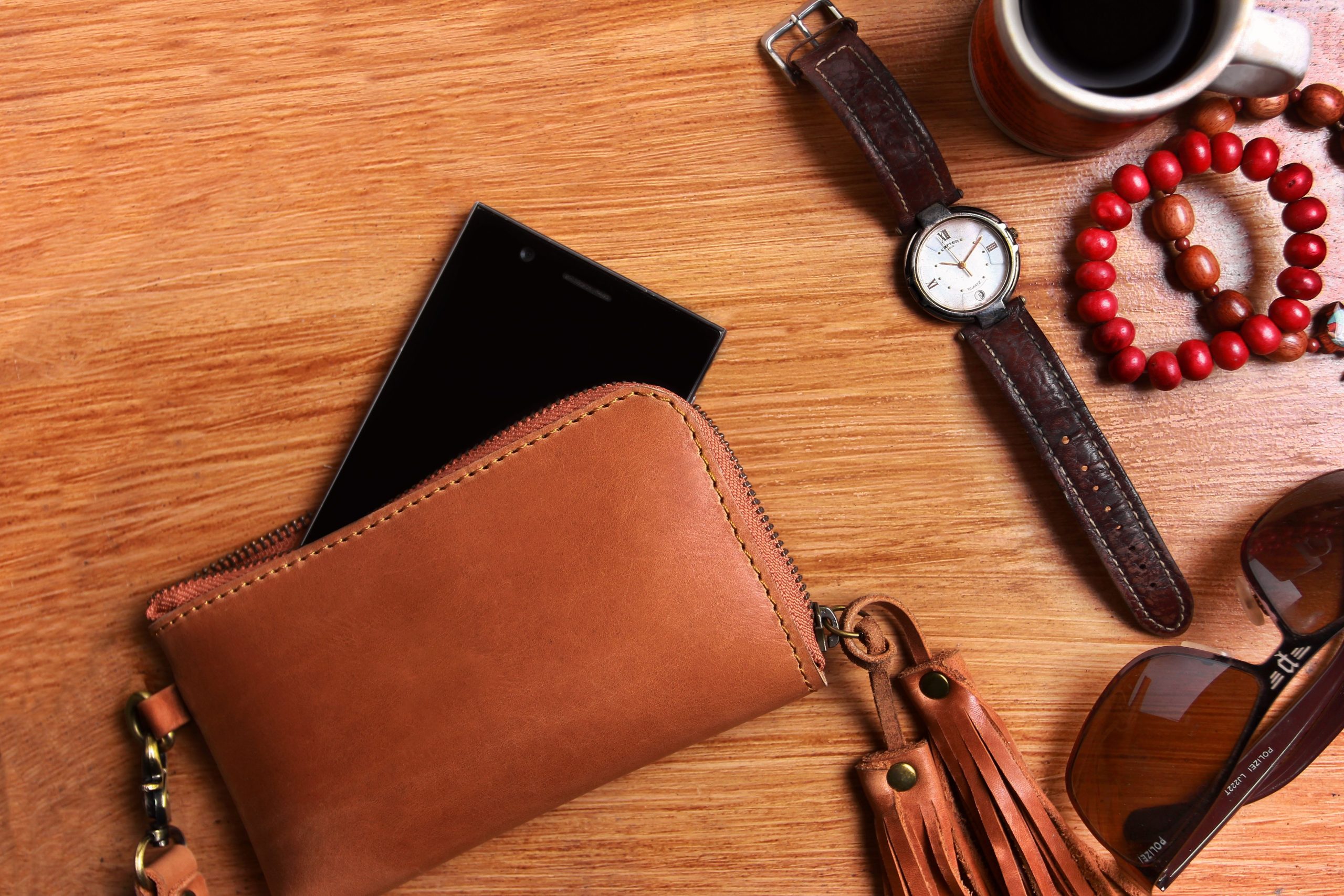 Tan leather zip-around wallet with a tassel and wrist strap, holding a phone, beside a leather-strap watch on a wooden table